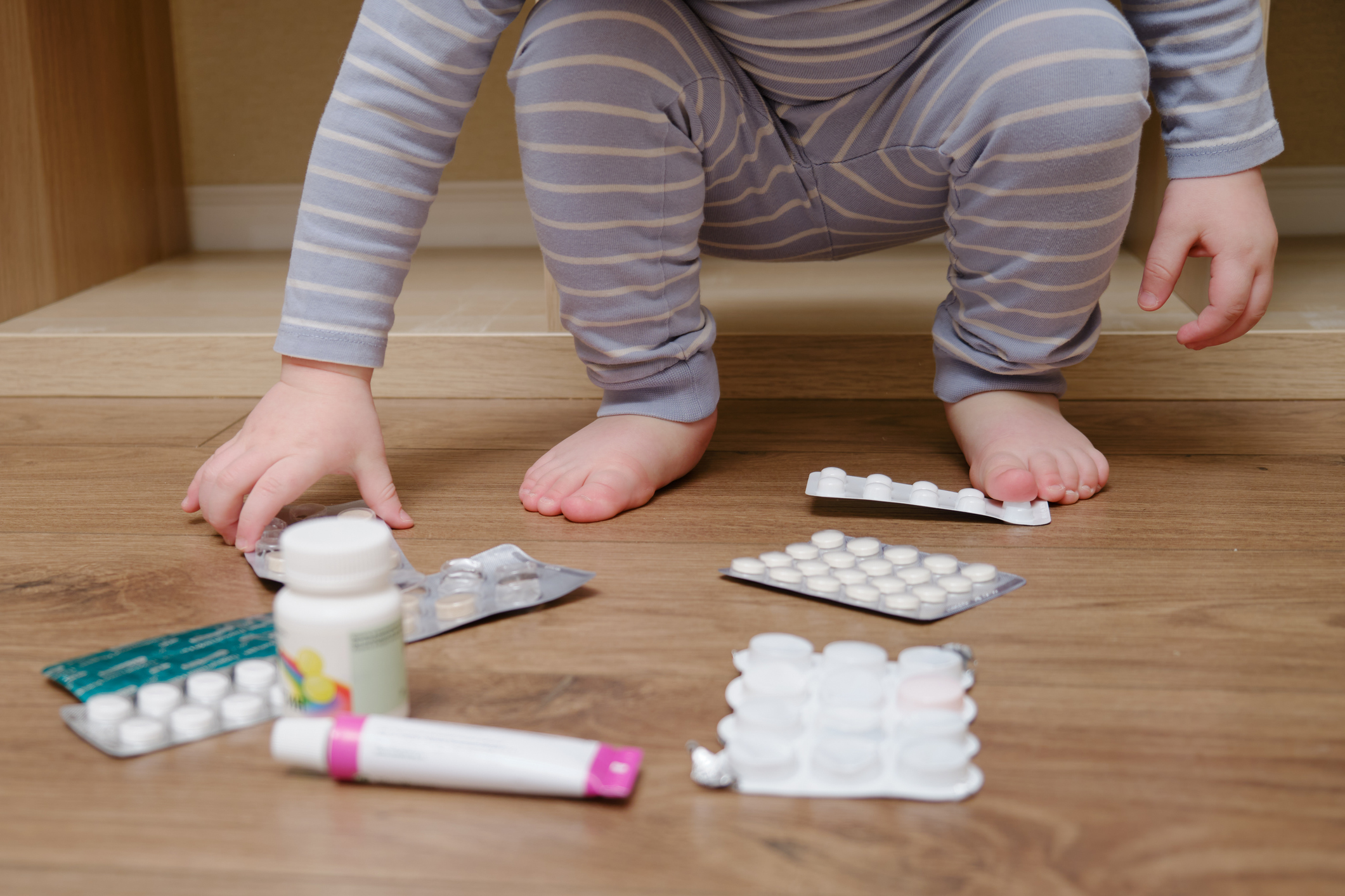 Child reaching down to grab a blister package of medicine from a pile of pill packages on the floor